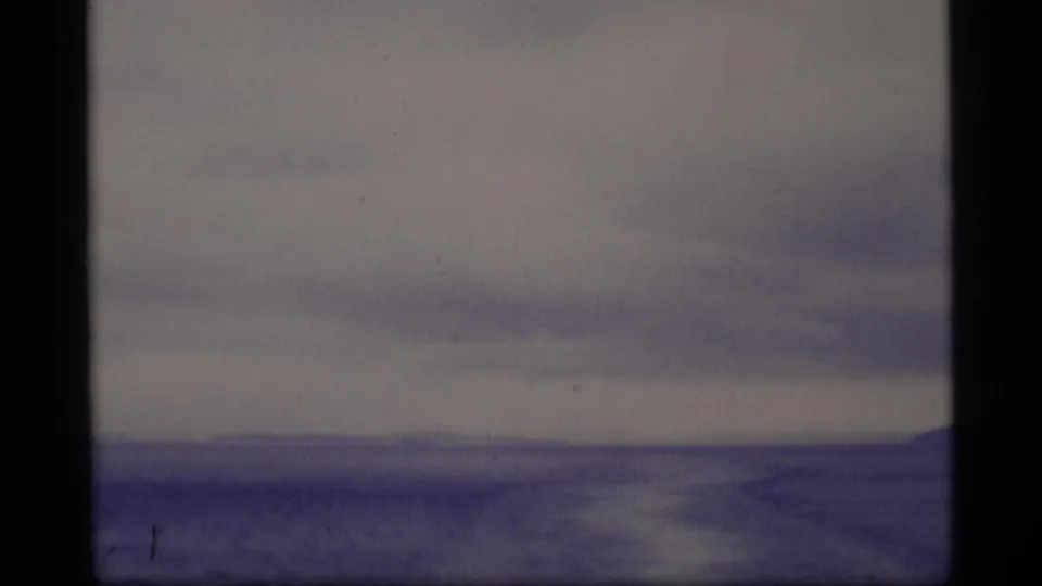 Blue-tinted 1977 Alaska 8mm frame showing open water or ice, far shoreline, and overcast sky through foreground framing.
