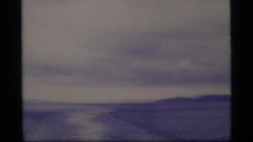 1977 Alaska 8mm view of low hills along a distant shore under layered clouds, with muted water or ice in the foreground.