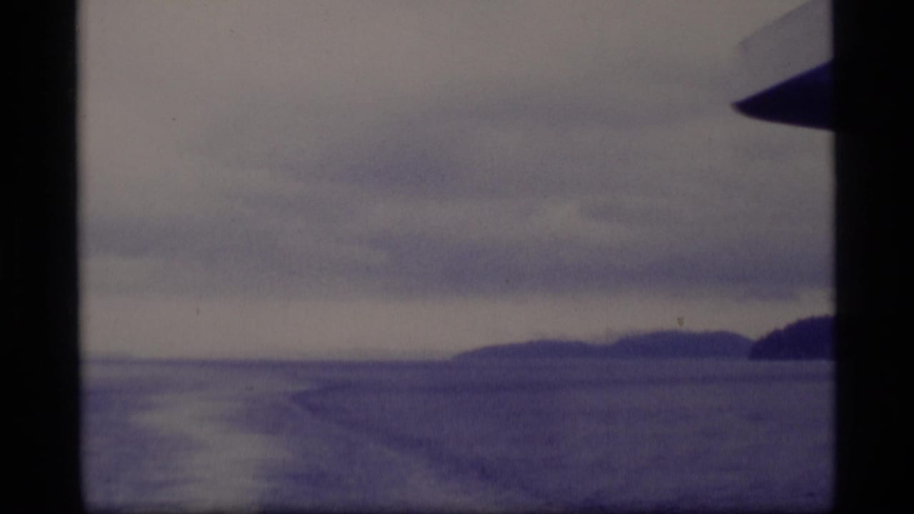1977 Alaska 8mm view from a vessel, with open water, distant landforms, cloudy sky, and dark vertical boat framing.