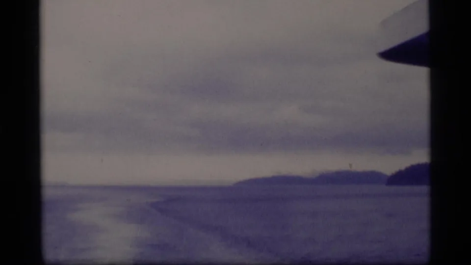 1977 Alaska 8mm view from a vessel, with open water, distant landforms, cloudy sky, and dark vertical boat framing.