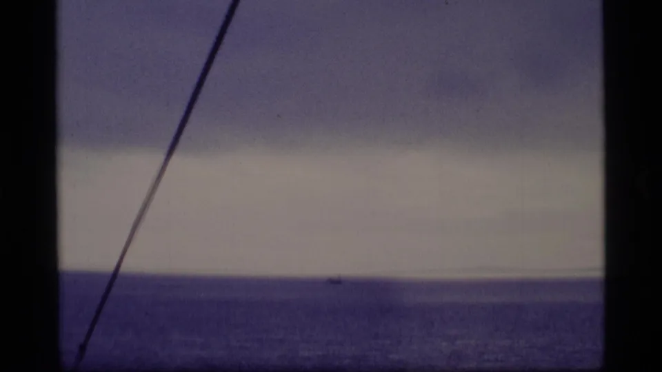 1977 Alaska 8mm frame of open sea and pale cloud band, with a diagonal mast or cable crossing the vessel-side view.