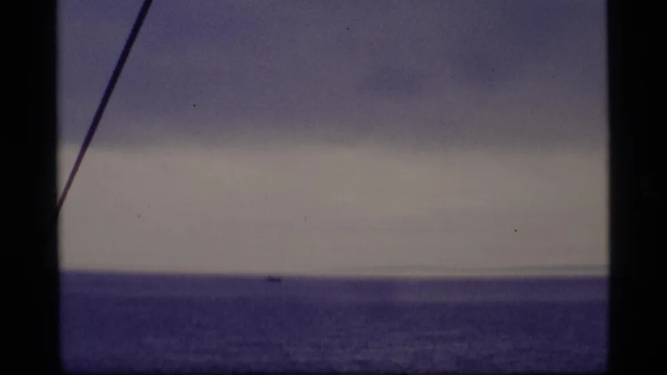 1977 Alaska 8mm vessel view of calm ocean, layered clouds, and a diagonal cable, with no people or structures visible.