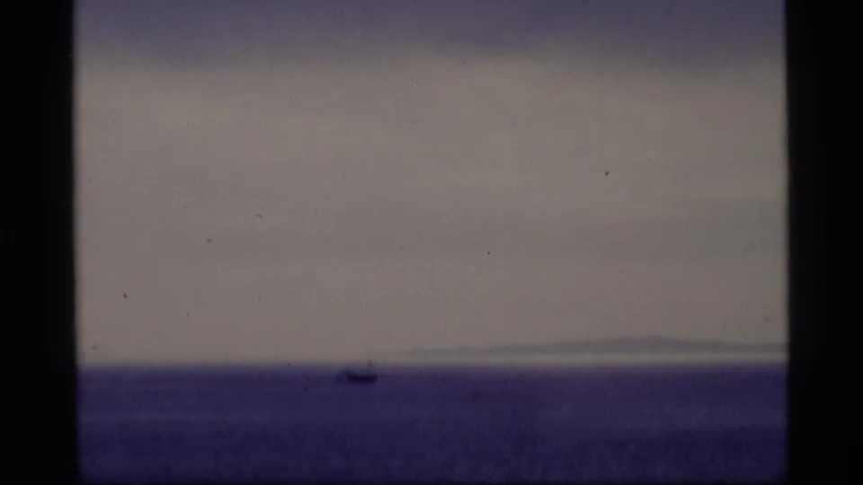 1977 Alaska 8mm frame showing a lone vessel as a dark silhouette against open water and a faint distant shoreline.