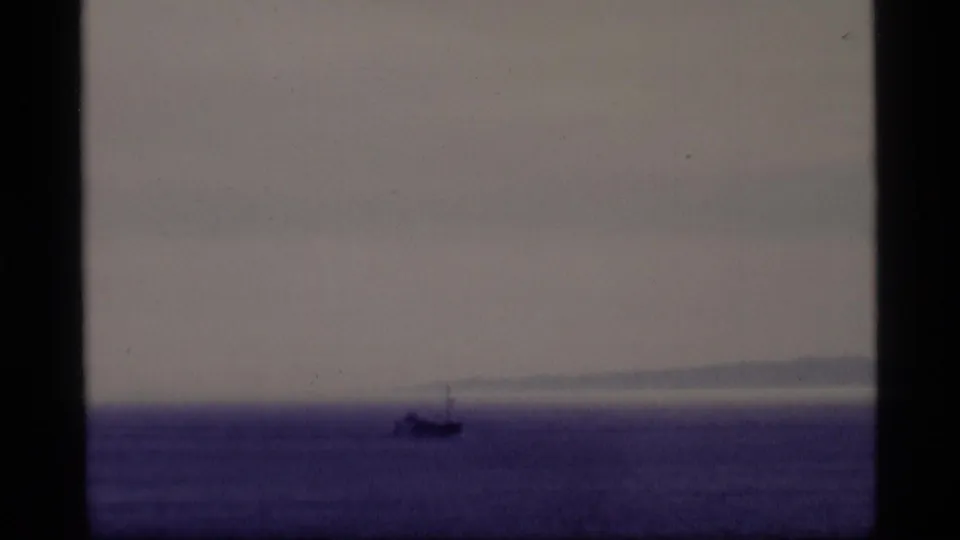 1977 Alaska 8mm frame with a small vessel low on the water and hazy coastal land visible beyond it.