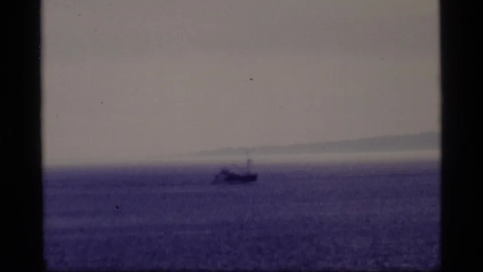 1977 Alaska 8mm frame showing a lone boat on purple-blue water with distant hills softened by haze.