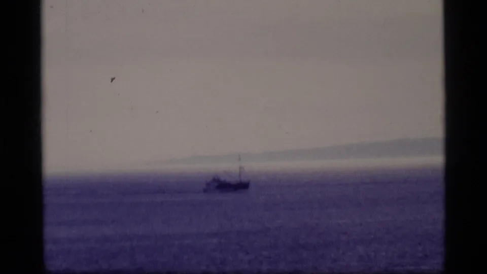 1977 Alaska 8mm frame showing a distant vessel on calm purple-tinged water with a faint landmass beyond.