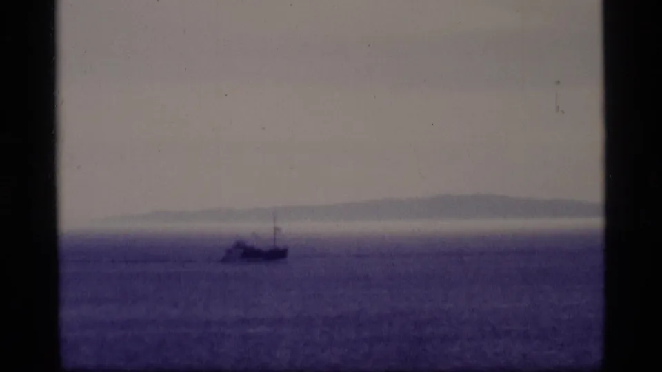 1977 Alaska home-movie frame with a small boat near the horizon and a soft, hazy shoreline behind it.