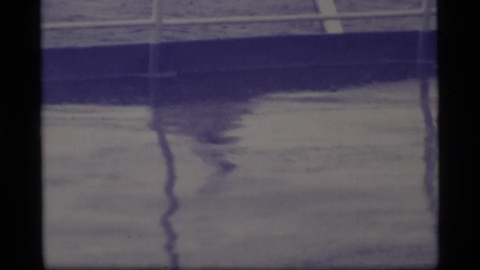 1977 Alaska 8mm frame looking down at a wet deck, metal railings, and dark reflected shapes on the surface.
