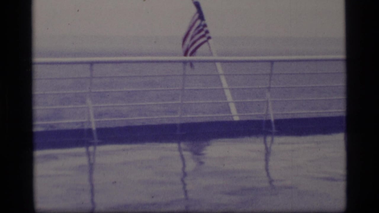 American flag flying from a ship deck in Alaska travel footage, with wet railings and open ocean visible through a blue-tinted 8mm frame.
