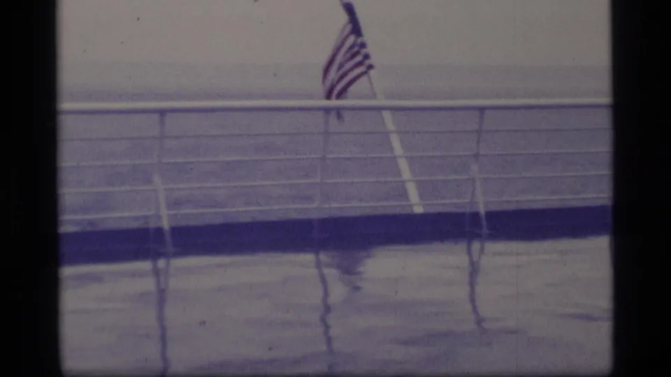 American flag flying from a ship deck in Alaska travel footage, with wet railings and open ocean visible through a blue-tinted 8mm frame.