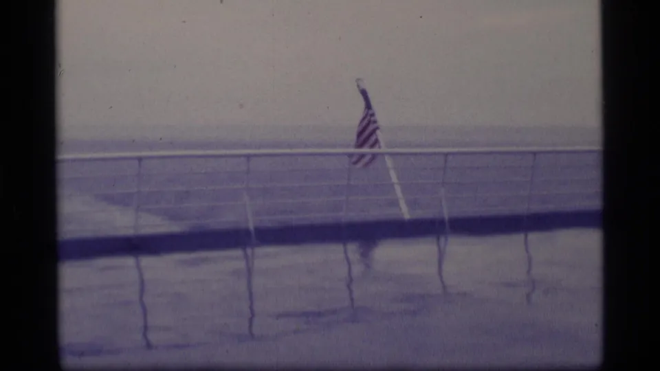 American flag on a slanted pole above a ship deck, with railings, ocean horizon, and overcast sky in a 1970s Alaska cruise context.