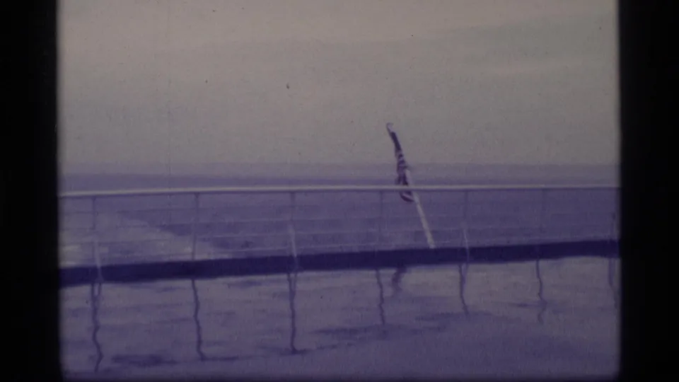 Blue-toned 8mm view of an American flag on a ship deck, with railing lines and wet deck reflections during a 1970s Alaska voyage.