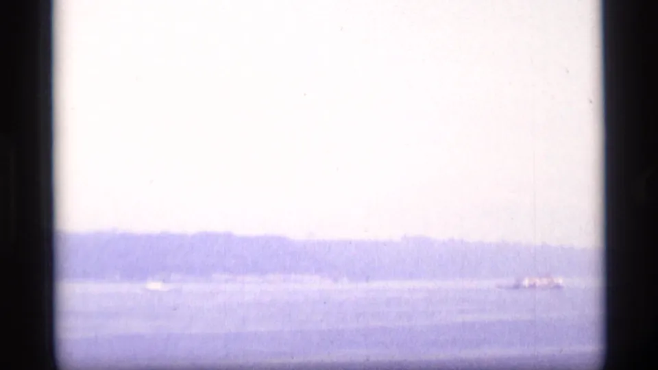 Distant Alaska waterfront view with a hazy shoreline and a small aircraft-like shape near the right edge, seen through blue-tinted 8mm film.