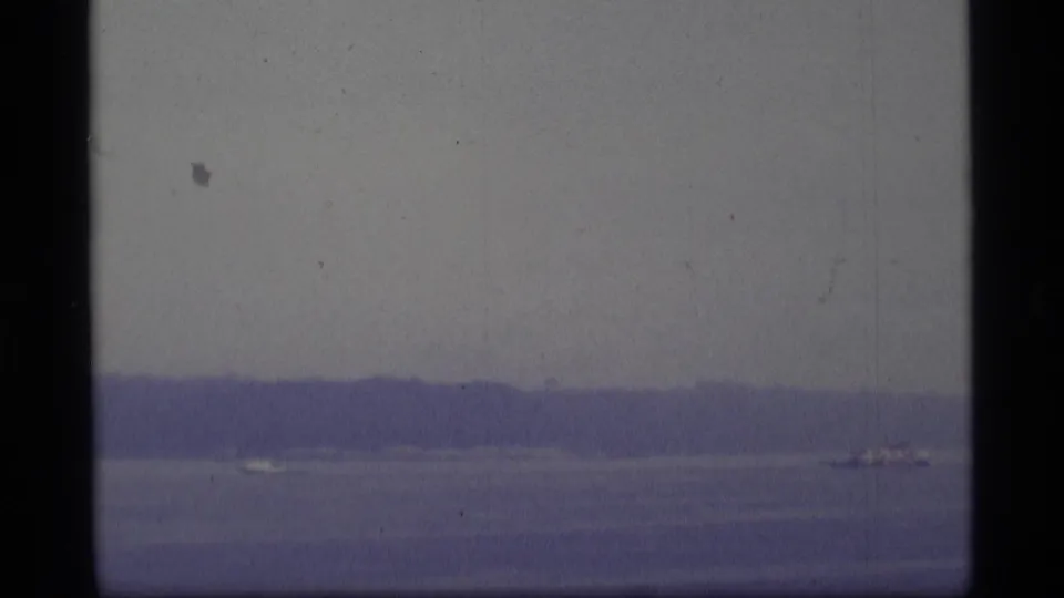 Wide 1977 Alaska water view with a low wooded shoreline and distant small aircraft shapes, softened by grain and blue-purple color shift.