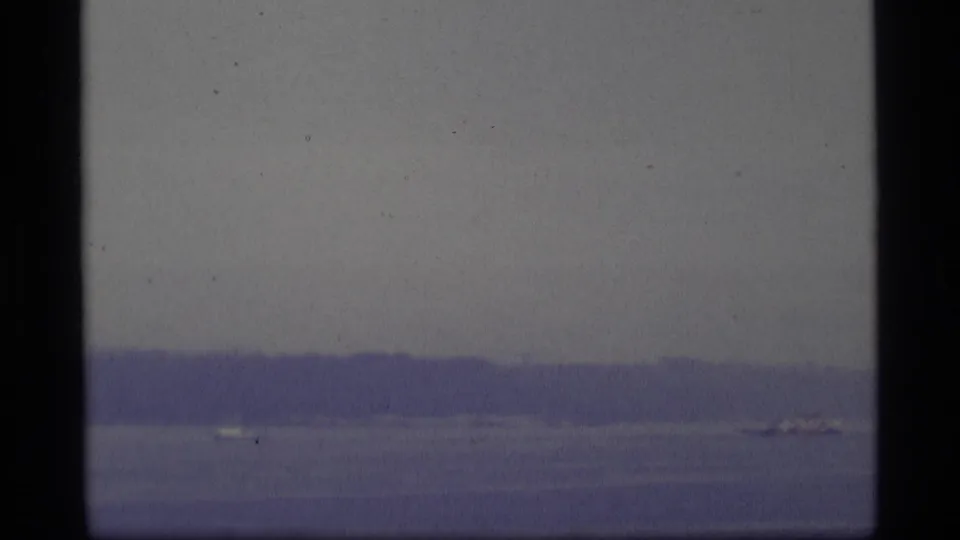 Distant Alaska shoreline and water with a small aircraft-like form near the right side, captured in soft blue-tinted 8mm home-movie footage.