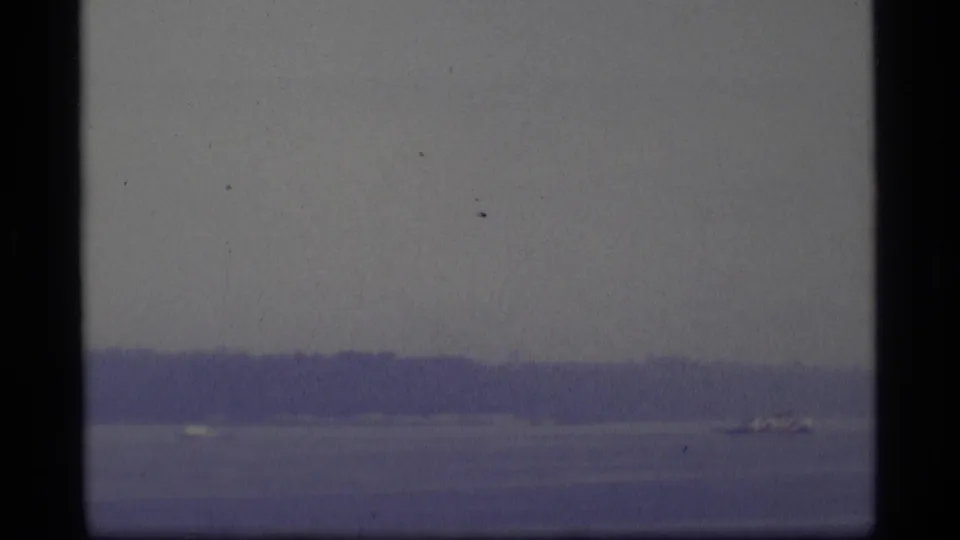 1977 Alaska 8mm frame showing a small distant boat crossing calm water below a hazy tree-lined shore.