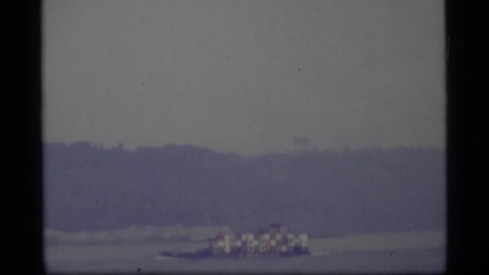 1977 Alaska home-movie frame with a small boat and checkered canopy moving across a hazy waterway.