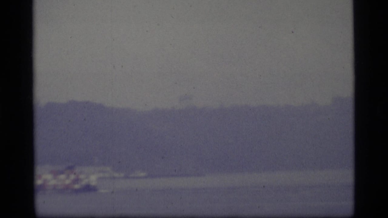 Soft 1977 8mm-style view of gray harbor water and a hazy distant shoreline, likely part of a Seattle waterfront sequence.