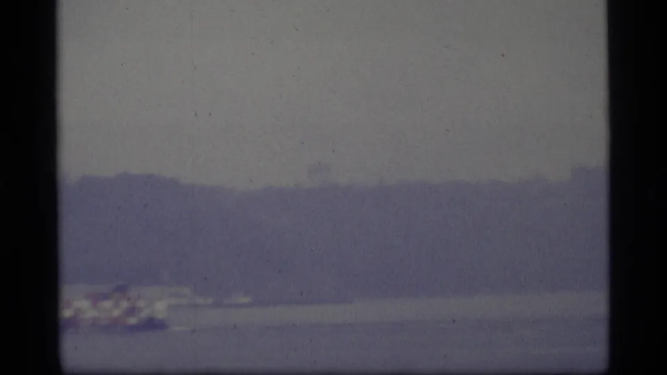 Soft 1977 8mm-style view of gray harbor water and a hazy distant shoreline, likely part of a Seattle waterfront sequence.