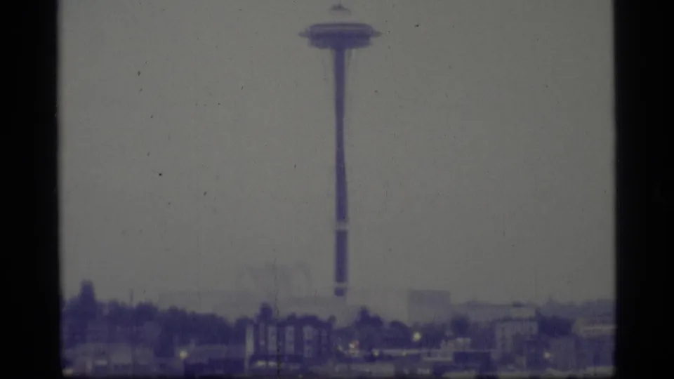 The Space Needle rises above low waterfront buildings in a fog-softened Seattle skyline view from 1977 home-movie footage.