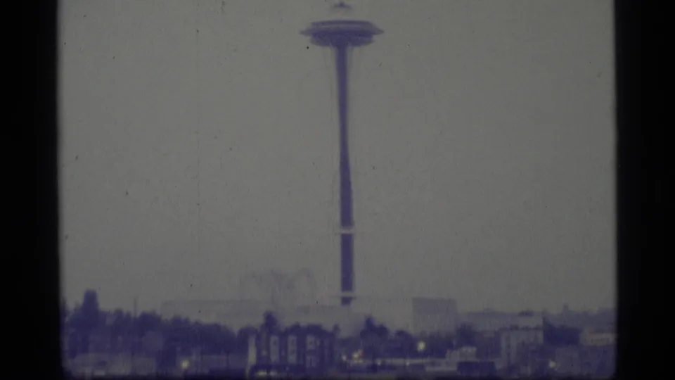 1977 8mm home-movie frame of a hazy urban skyline, with a slender observation tower centered above low buildings and trees.