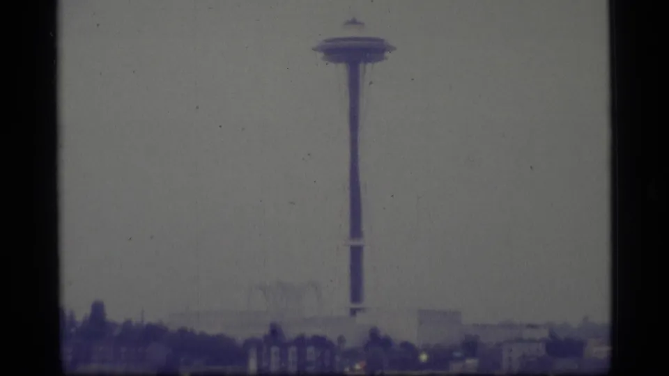 1977 8mm frame showing a distant observation tower over a soft, gray city view with low buildings and trees below.