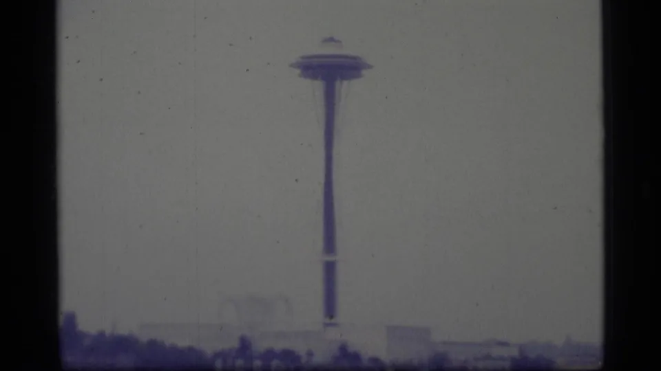 1977 8mm home-movie frame with the observation tower enlarged in the center and a faint urban skyline at the bottom edge.