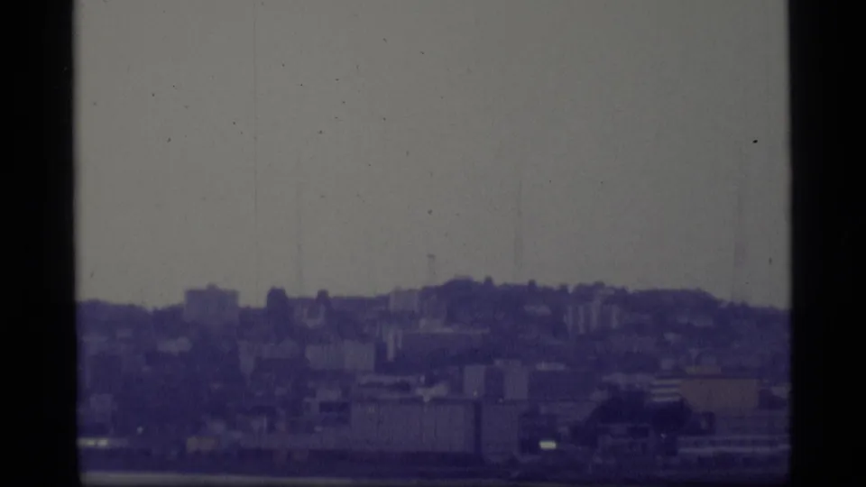 1977 8mm frame shifting to a broader city view, with hillside buildings, waterfront-like structures, and faint tower forms in haze.