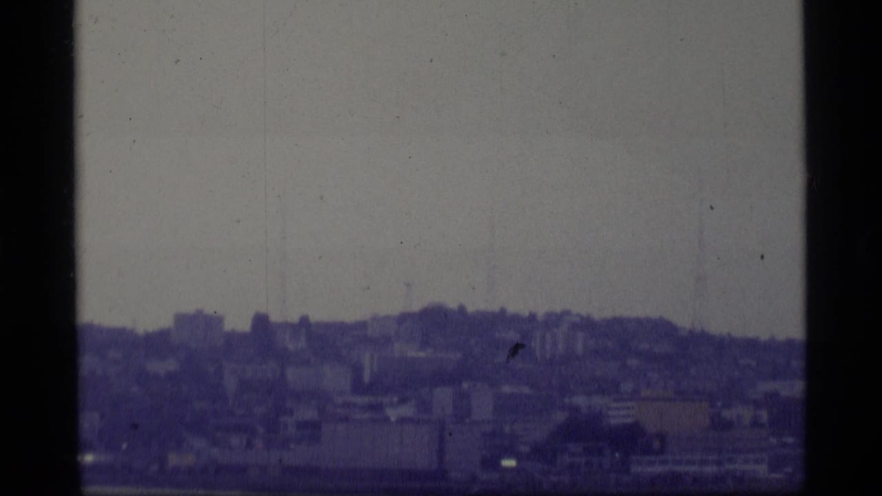 1977 Alaska home-movie frame showing a soft, hazy urban skyline across water with hills behind it.