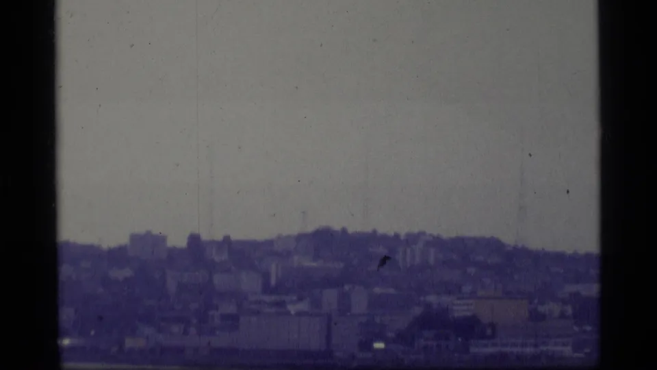 1977 Alaska home-movie frame showing a soft, hazy urban skyline across water with hills behind it.