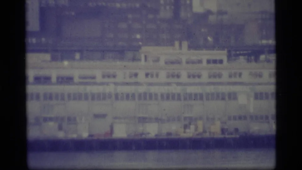 Vintage 8mm frame showing a large pale waterfront vessel or industrial structure below a blurred city backdrop.