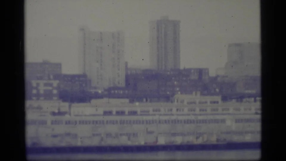 1977 Alaska waterfront cityscape with tall buildings in the distance and a large pale structure along the water.