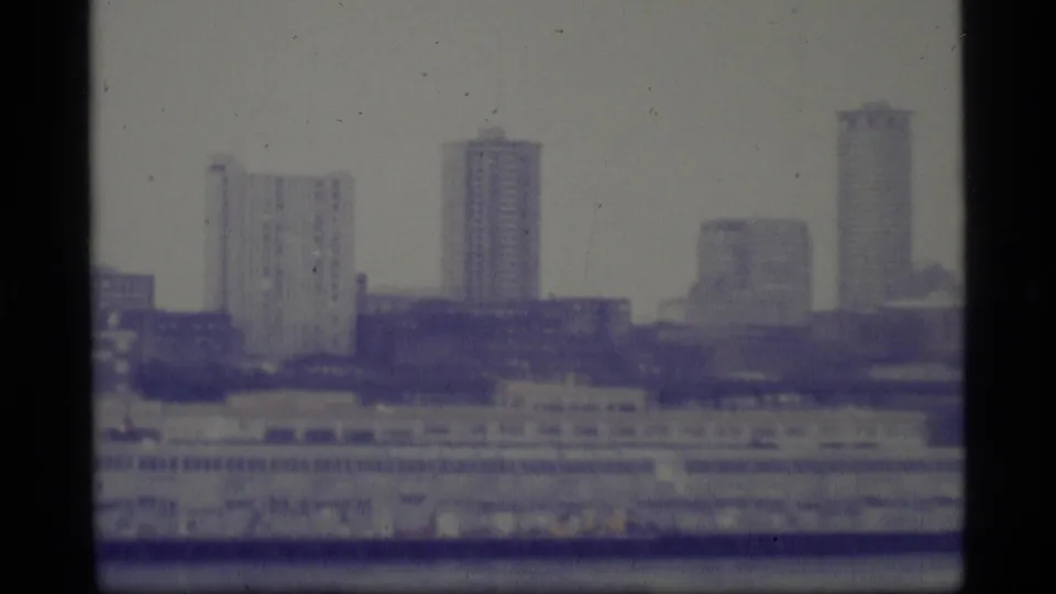 1977 Alaska home-movie frame showing a hazy urban skyline and waterfront seen from across a river or harbor.