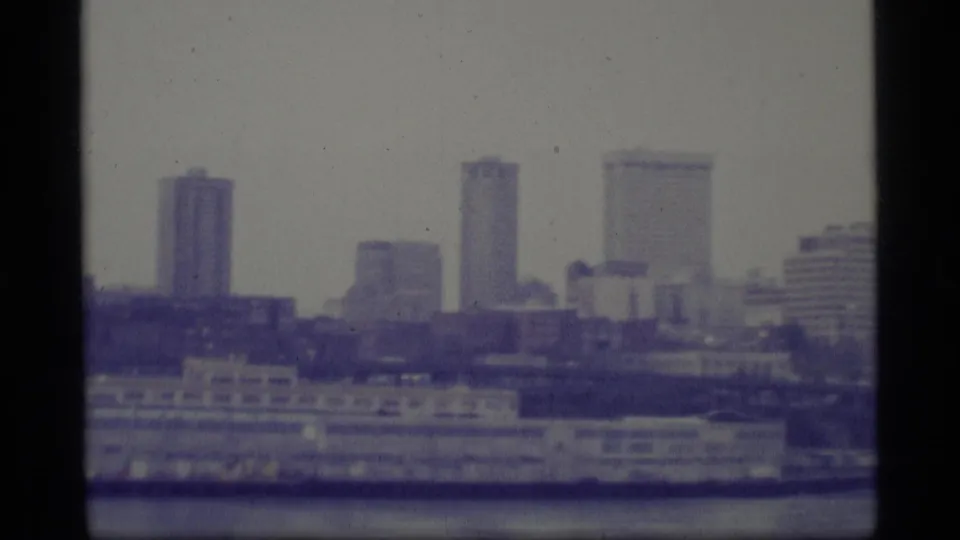 Blue-tinted 1977 Alaska frame with mid-century high-rises and lower waterfront structures across the water.