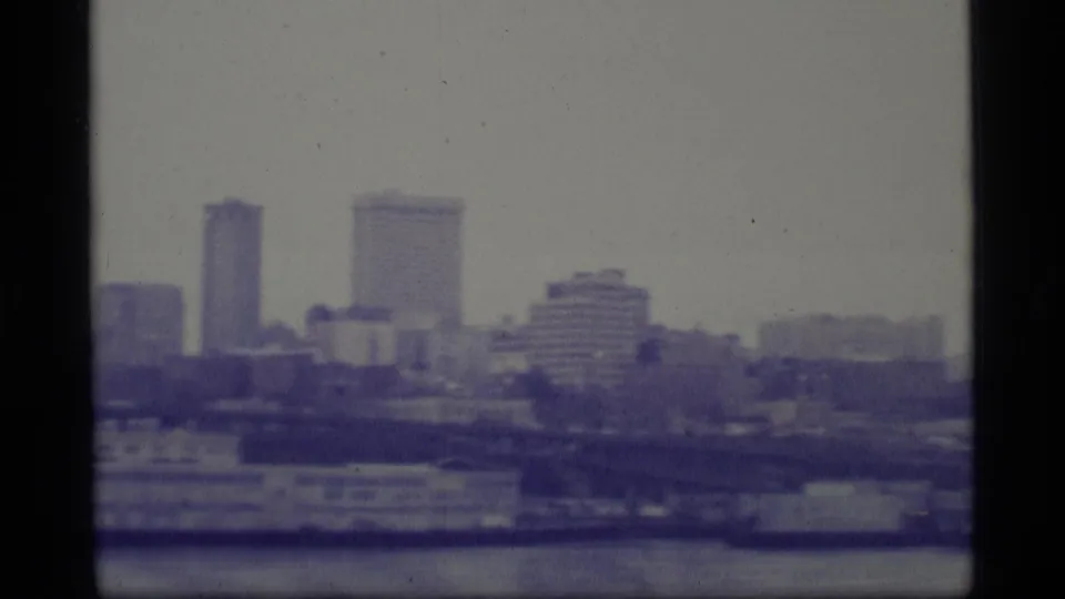 Archival 8mm frame of an Alaska waterfront skyline, with tall buildings, low structures, and water in the foreground.