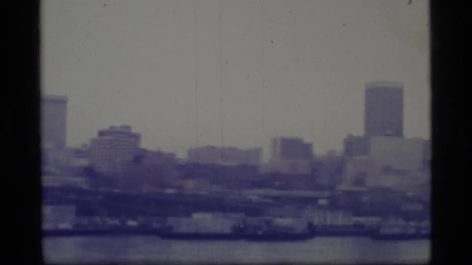 Late pan frame from a 1977 Alaska home movie showing distant waterfront buildings and a tall tower near the right edge.