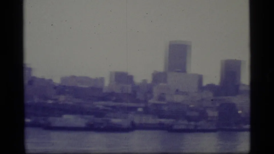 1977 8mm Alaska footage appears to show a distant city skyline across water under an overcast sky.
