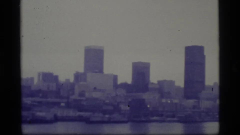A hazy 1977 8mm view of an Alaska city skyline across water, framed by visible film-strip edges.