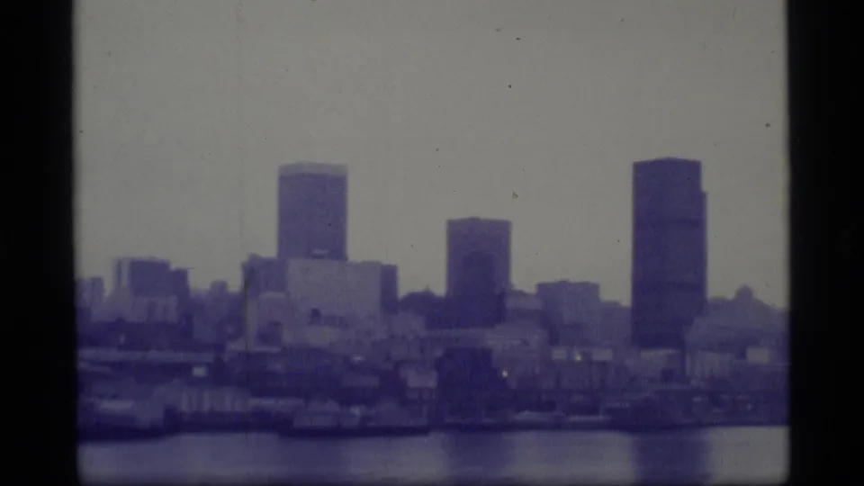 1977 8mm Alaska home-movie frame showing towers and low waterfront structures across a body of water.