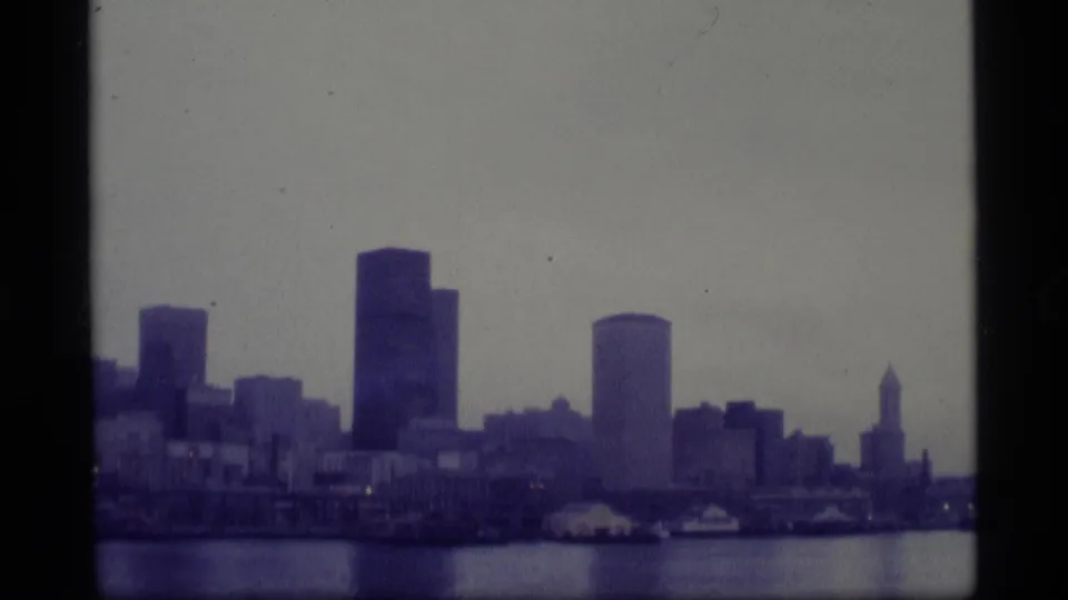 A wider 1977 8mm Alaska skyline view across water, with waterfront structures and distant high-rises.