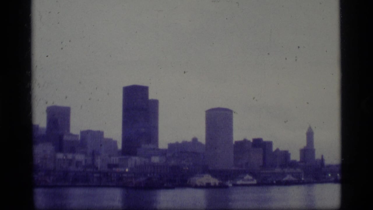 1977 Alaska 8mm frame showing a soft, distant waterfront skyline across harbor water, with high-rise buildings and visible film gate edges.