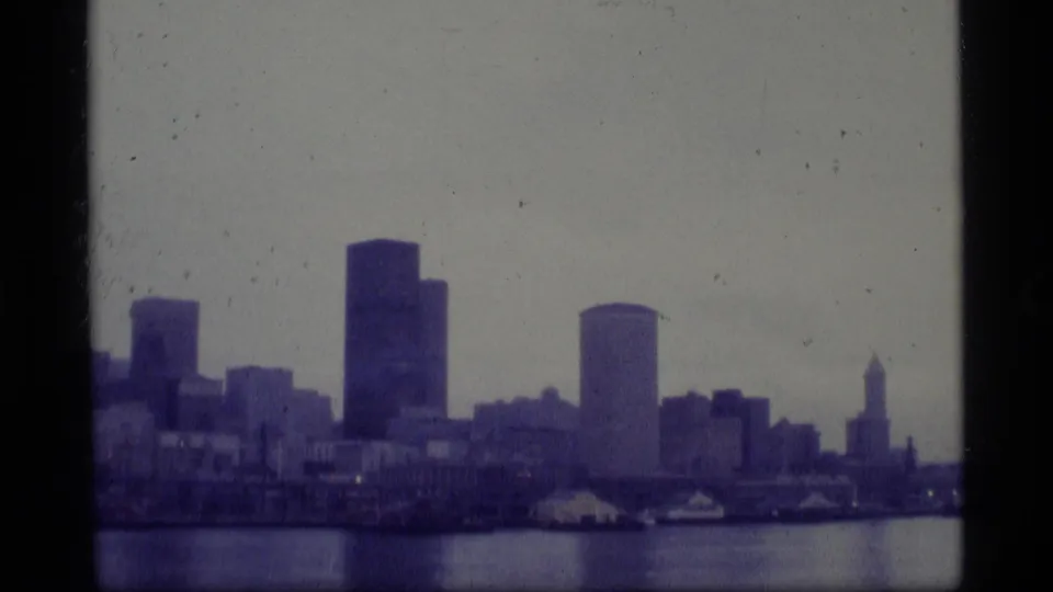 1977 Alaska 8mm frame showing a soft, distant waterfront skyline across harbor water, with high-rise buildings and visible film gate edges.