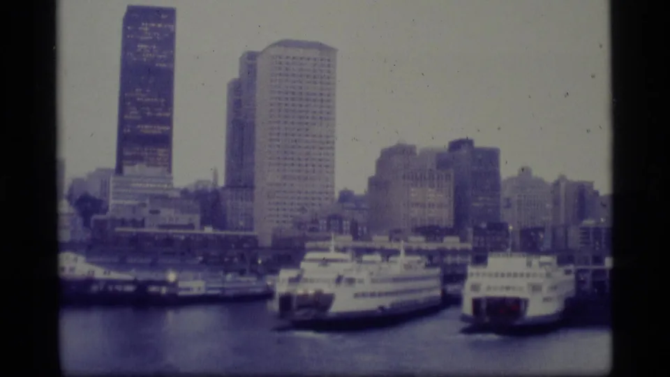 1977 Alaska 8mm frame showing ferries or cruise vessels at an urban waterfront, with tall office buildings rising behind the docks.