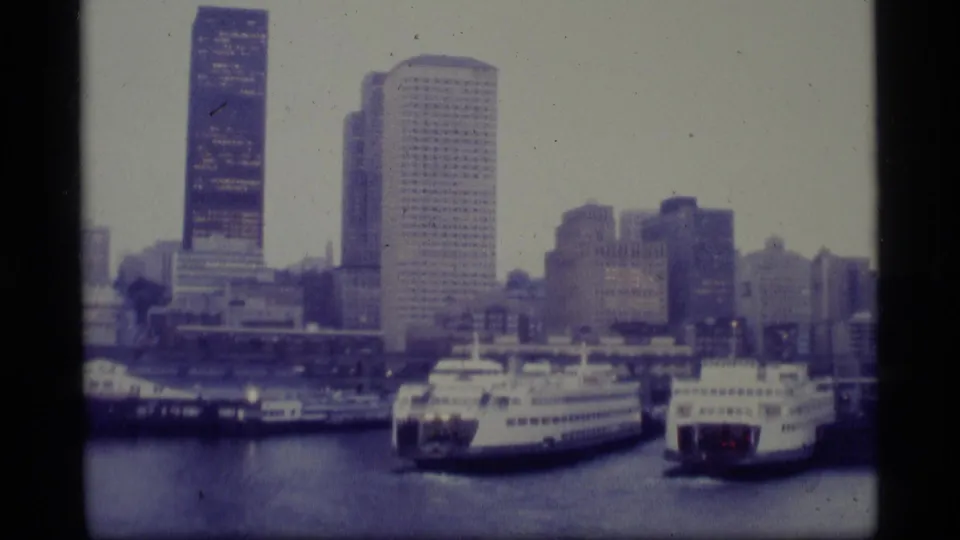 1977 Alaska 8mm frame with two docked ferries or cruise liners in the foreground and an urban harbor skyline behind them.
