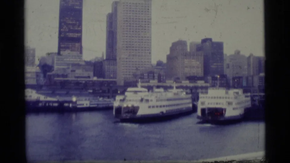 1977 Alaska 8mm frame showing docked passenger ferries at a city harbor, with waterfront structures and tall buildings behind them.