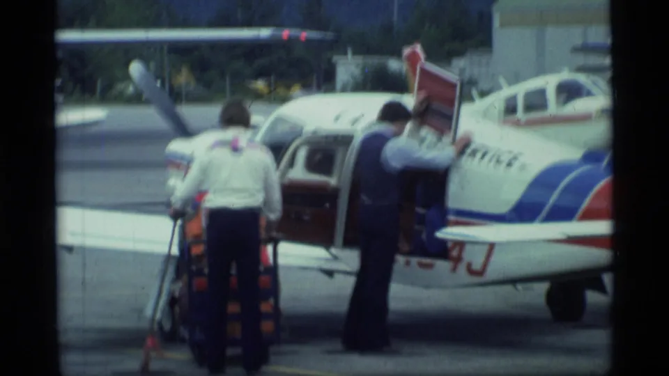 In 1977 Alaska footage, two men work beside a white small aircraft with red and blue stripes and an open cabin door on an airport ramp.