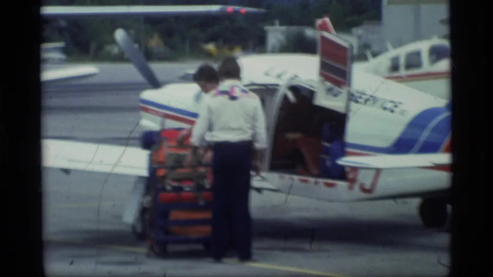 Two men stand at the open door of a small white airplane with red and blue striping, with cargo or equipment stacked on a hand cart nearby.
