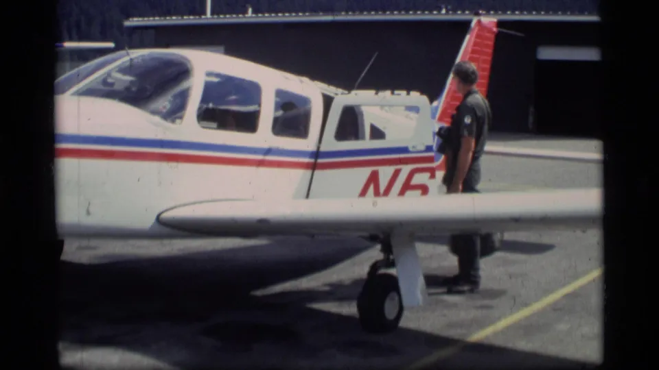 A man stands by the tail and open side area of a small white aircraft with red and blue stripes on an Alaska airport ramp in 1977 footage.