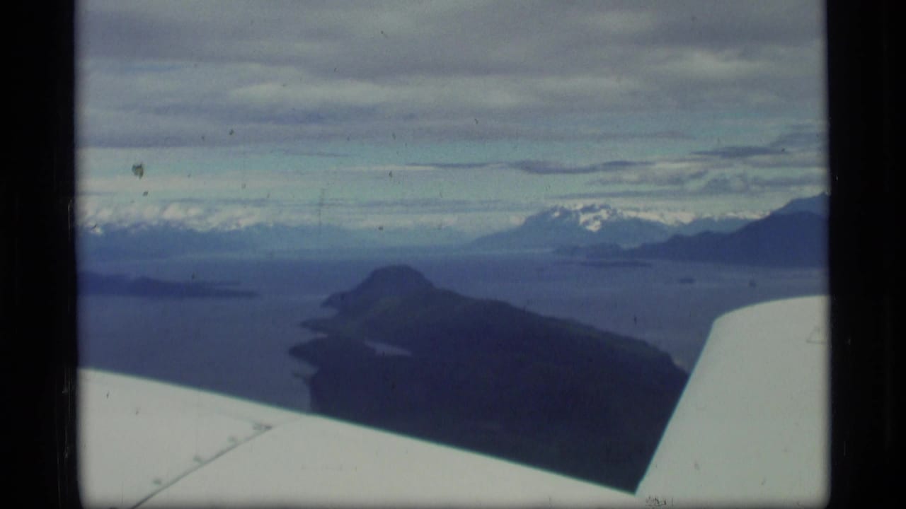 1977 Alaska 8mm aerial view from an aircraft window over coastal water, dark landforms, distant snow-capped mountains, and cloud cover.