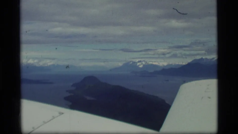 1977 Alaska aerial home-movie frame showing the aircraft wing edge above water, dark island terrain, distant snowy peaks, and a heavy sky.